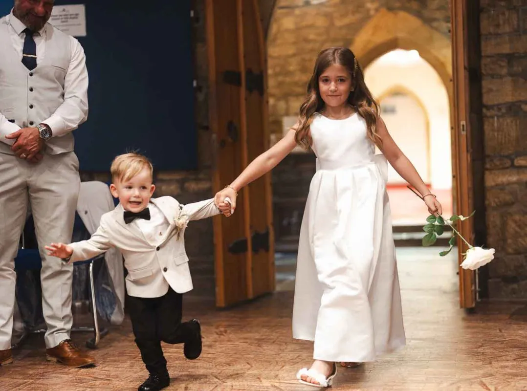 A young flower girl and page boy happily walk hand-in-hand down the aisle through a stone archway during a church wedding ceremony.