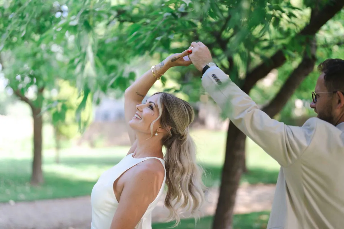 A bride and groom share a joyful dance in a lush garden, surrounded by trees filtering the soft summer sunlight.