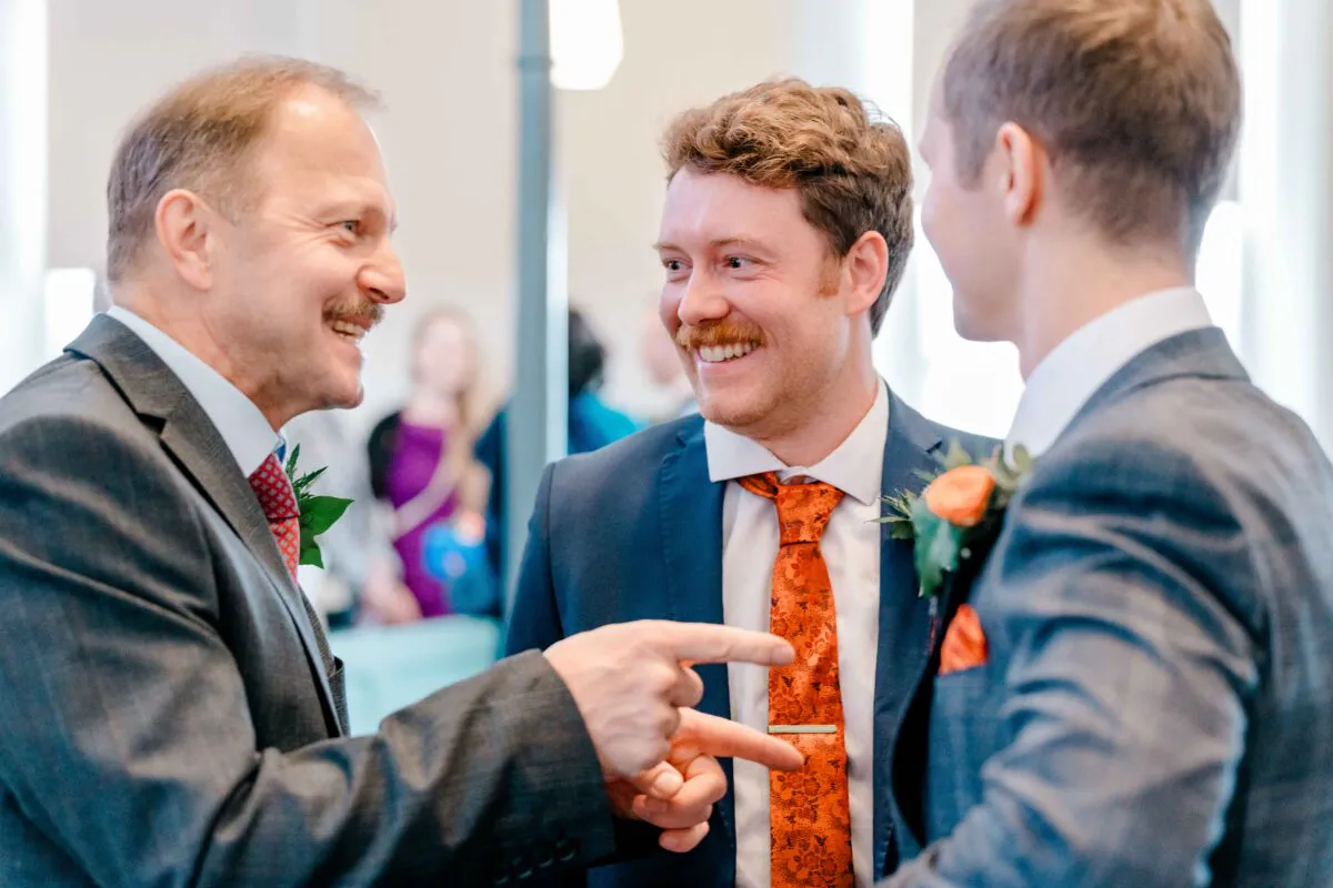 Three smiling men in tailored suits are chatting and laughing together at a wedding reception.