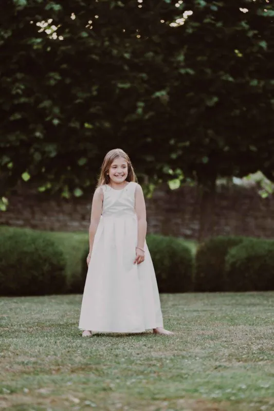 A smiling flower girl in a white dress stands barefoot on a lawn in a garden setting, illuminated by soft dappled sunlight.