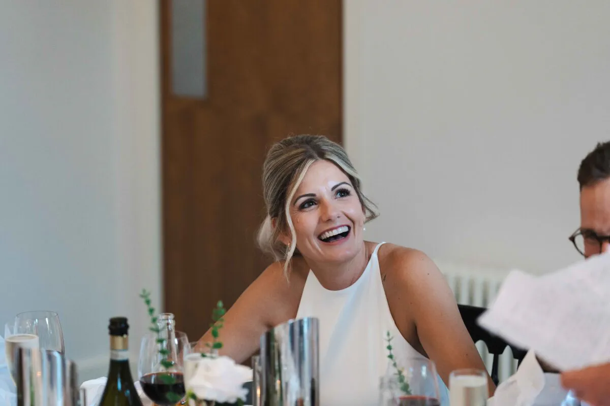 A smiling blonde woman laughs joyfully during the wedding speeches, seated at a reception table with wine glasses and tableware.