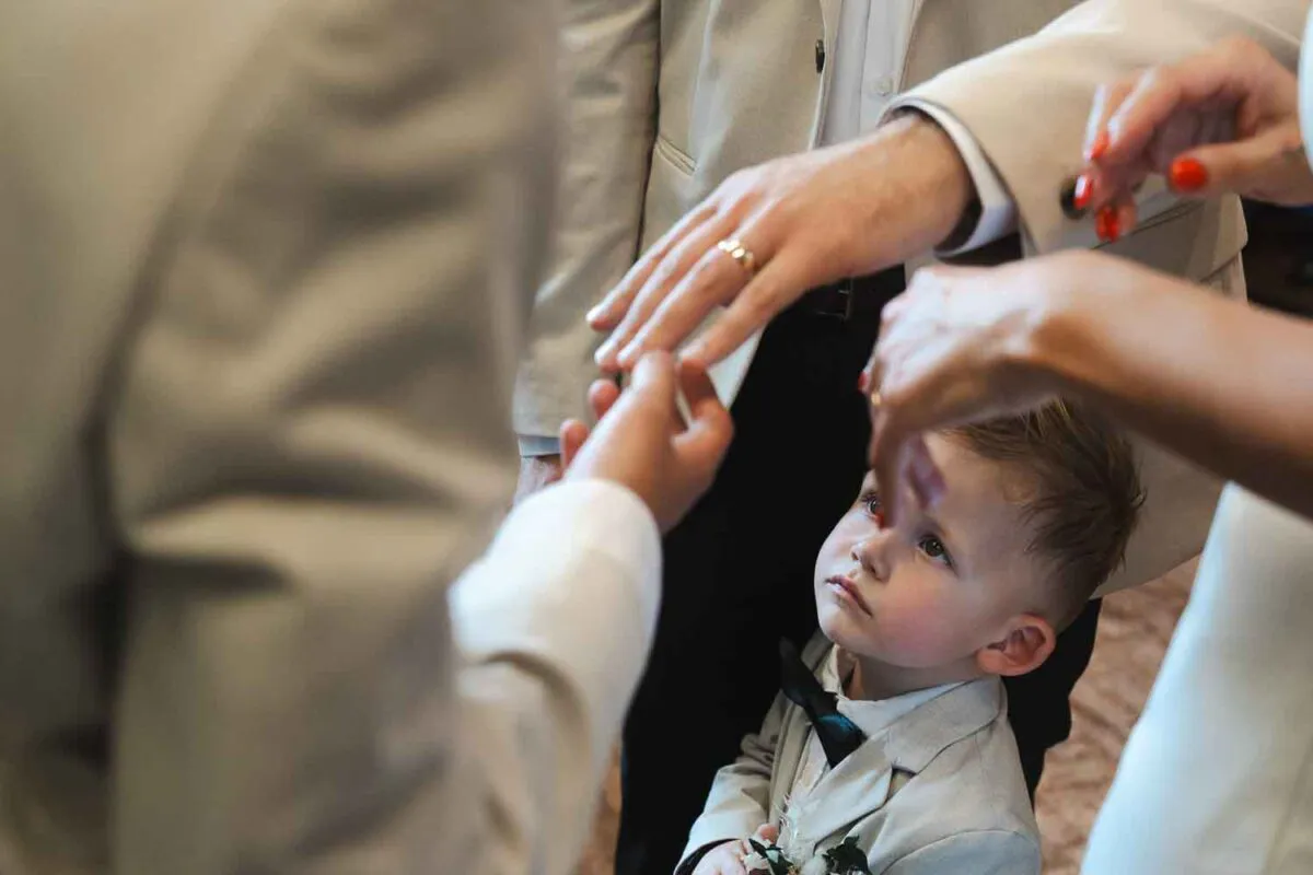 A young page boy wearing a light grey suit and bow tie looks up curiously while family members exchange wedding rings during the ceremony.