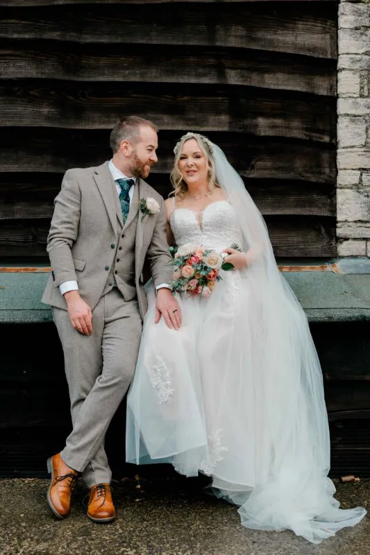 A smiling bride holding a colourful bouquet and wearing a tulle gown sits beside her groom in a tweed suit in front of a rustic wooden barn.