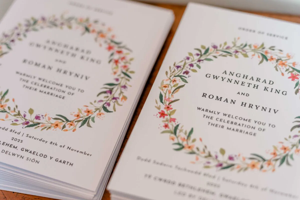 Close-up of stacked order of service booklets featuring a delicate watercolour floral wreath design framing the couple's names.