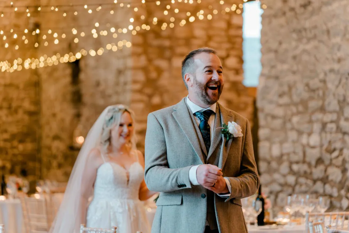 A groom in a grey suit laughs joyfully during speeches inside a rustic stone barn adorned with twinkling fairy lights, while the bride smiles in the soft-focus background.