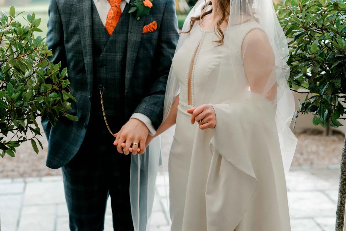 A newlywed couple holding hands outdoors, with the groom wearing a checked tweed suit and the bride in a crepe gown with pearl neckline detailing.