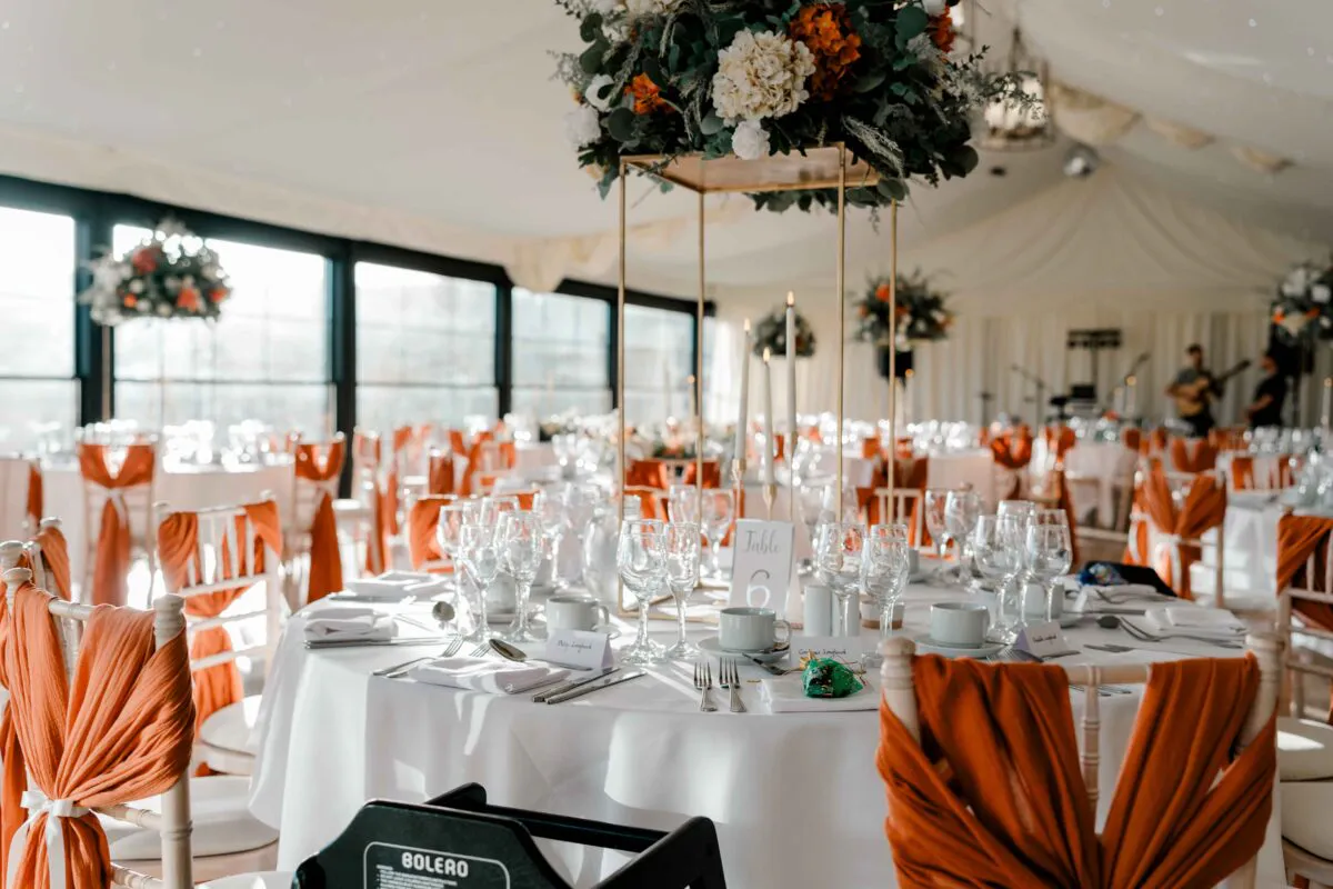 An elegantly styled round wedding table inside a bright marquee features a tall floral centrepiece stand with burnt orange blooms, taper candles, and matching orange chair sashes.