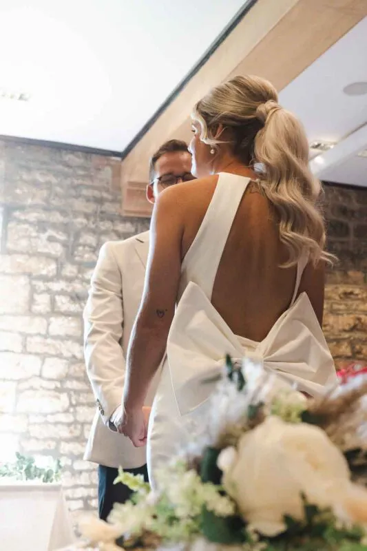 An over-the-shoulder view of a bride with a high ponytail and an oversized bow on her satin dress standing opposite the groom during their indoor ceremony.