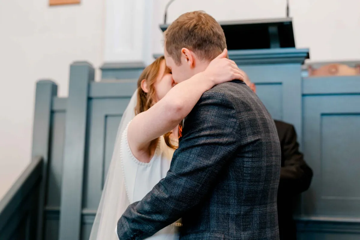 The bride and groom share their first kiss at the altar during a traditional church ceremony, with the groom wearing a grey checked suit.