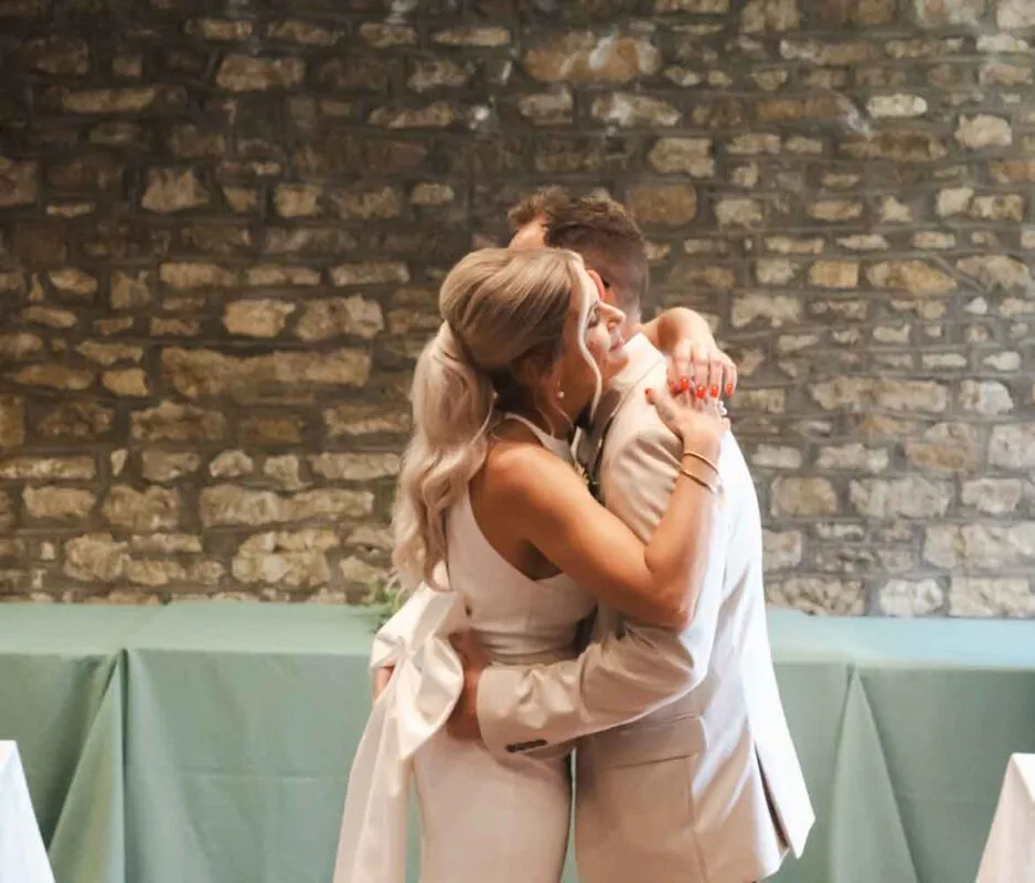 A newly married couple embraces tenderly in front of a rustic exposed stone wall during their wedding celebration.