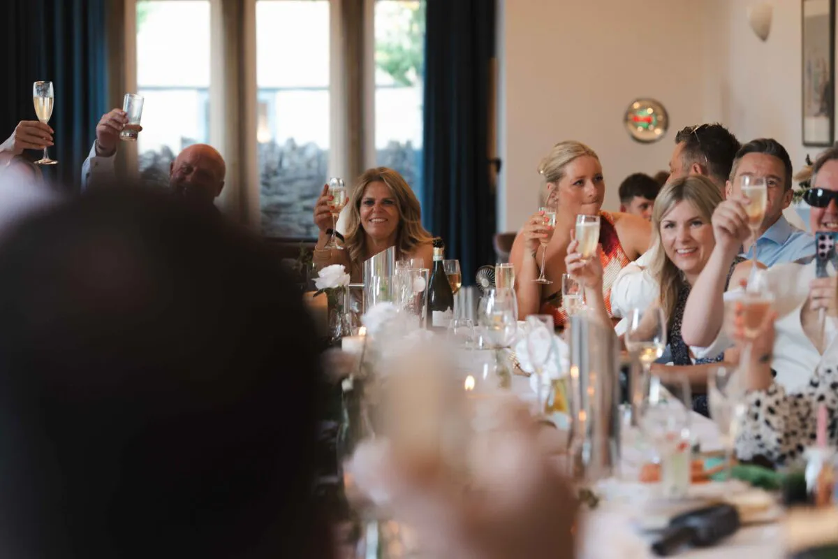 Wedding guests raise their glasses in a toast during the speeches, laughing and smiling around a beautifully set banquet table.