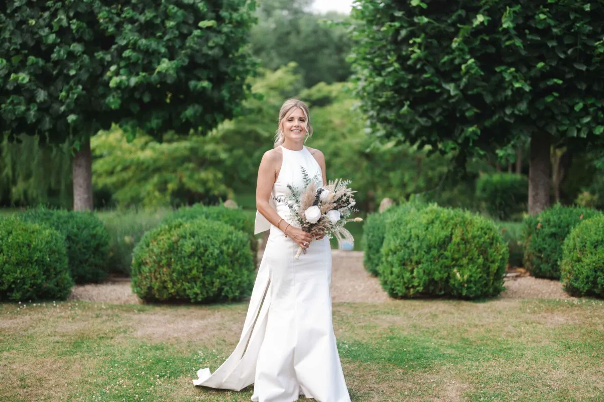 A smiling bride wearing a modern halterneck crepe wedding dress with a high slit holds an unstructured bouquet filled with roses and dried grasses in a manicured garden setting.