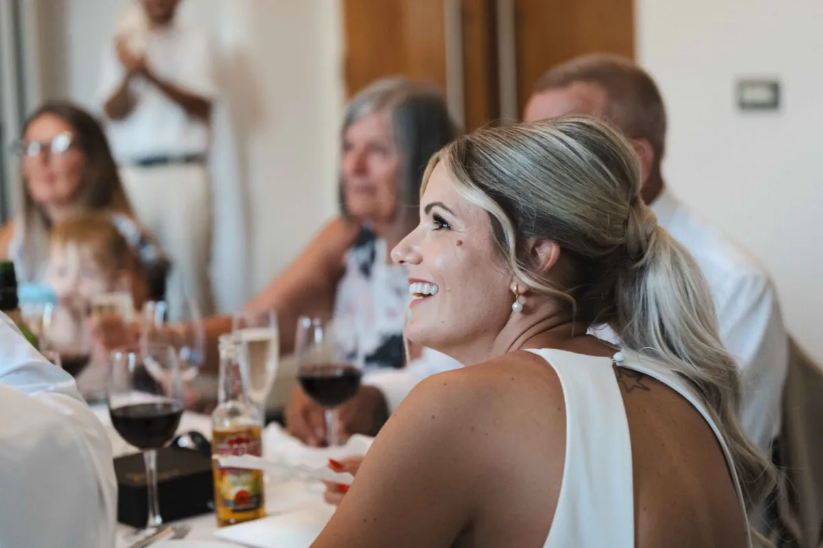 A guest at the wedding breakfast smiles and laughs joyfully while seated at a table during speeches, wearing a white dress and her blonde hair styled in a low ponytail.