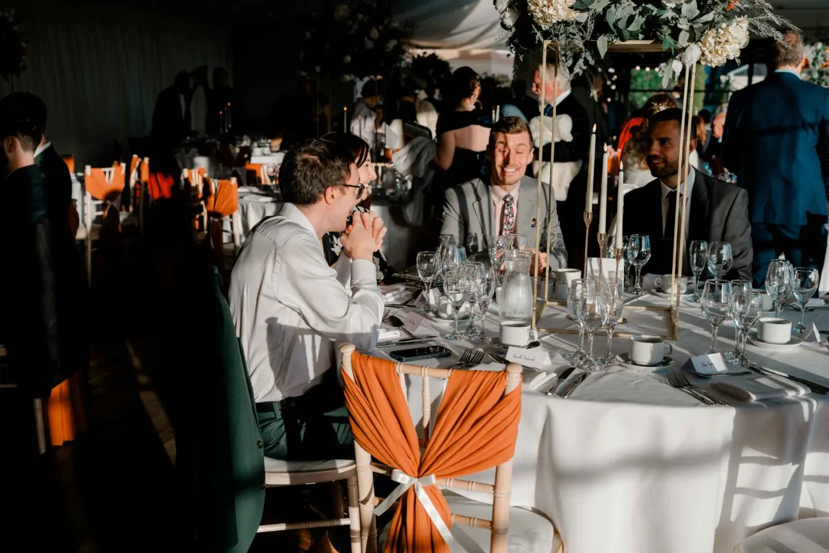 Wedding guests enjoy animated conversation and laughter around a sunlit banquet table adorned with elegant floral centerpieces and tapered candles during the reception.