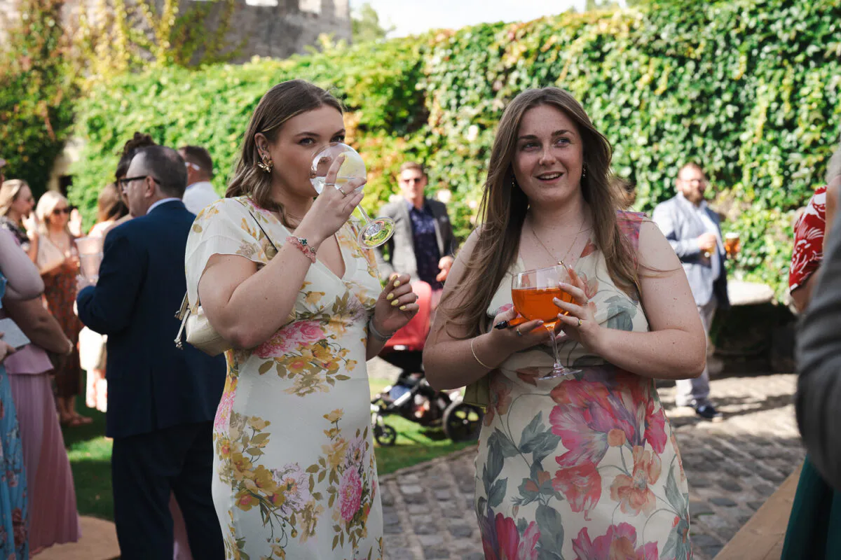 Two female wedding guests in floral dresses enjoy cocktails during an outdoor drinks reception in a cobbled courtyard.