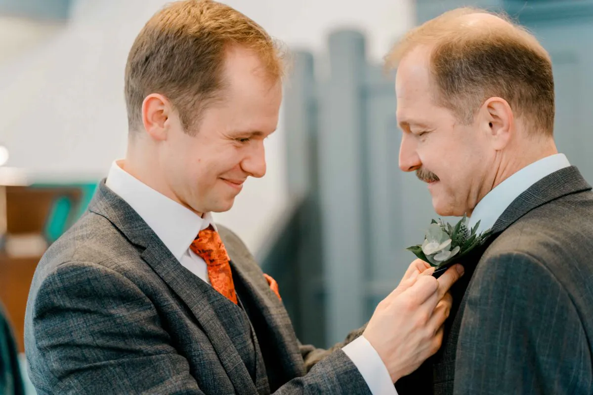 A groom in a grey suit and patterned orange tie carefully pins a floral buttonhole onto another man's lapel during wedding preparations.