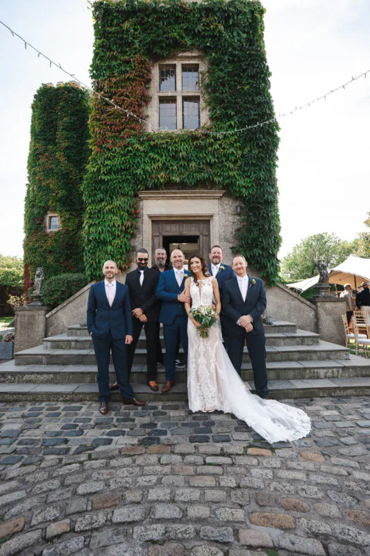 A smiling bride and groom pose with their groomsmen on stone steps outside an ivy-covered stone tower with a cobbled courtyard.