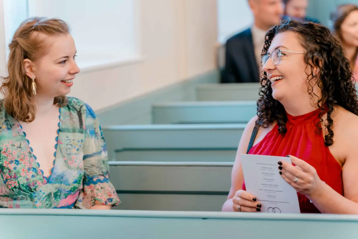 Two wedding guests sitting in pale green church pews smile and chat happily while holding an order of service.