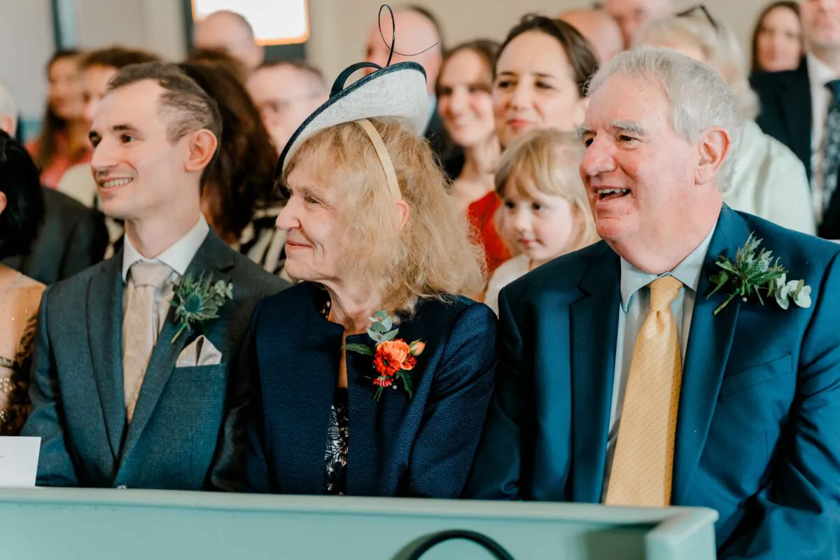 Smiling wedding guests seated on a pew during the ceremony, including older relatives wearing suits and a fascinator.