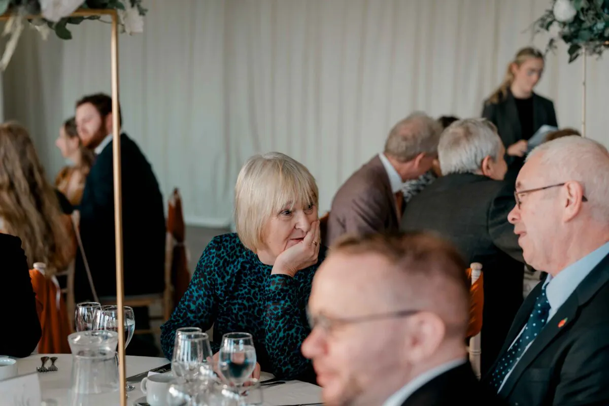 Wedding guests converse and mingle during the wedding breakfast seated around a table set with glassware and floral arrangements.