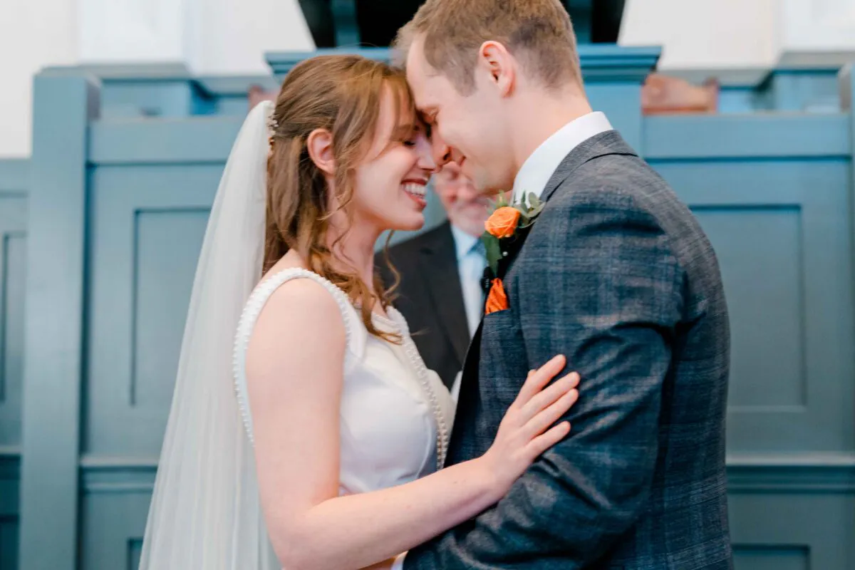 A newlywed bride and groom smile joyfully with their foreheads touching during an indoor wedding ceremony.