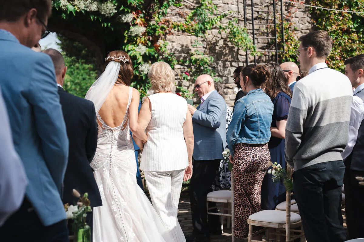 A bride walks down the outdoor ceremony aisle arm-in-arm with a guest, wearing a lace gown with an open back and a veil, watched by seated guests.
