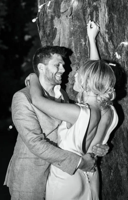 A newlywed couple embraces and smiles lovingly against a tree trunk outdoors at night, captured in a monochrome photographic style.