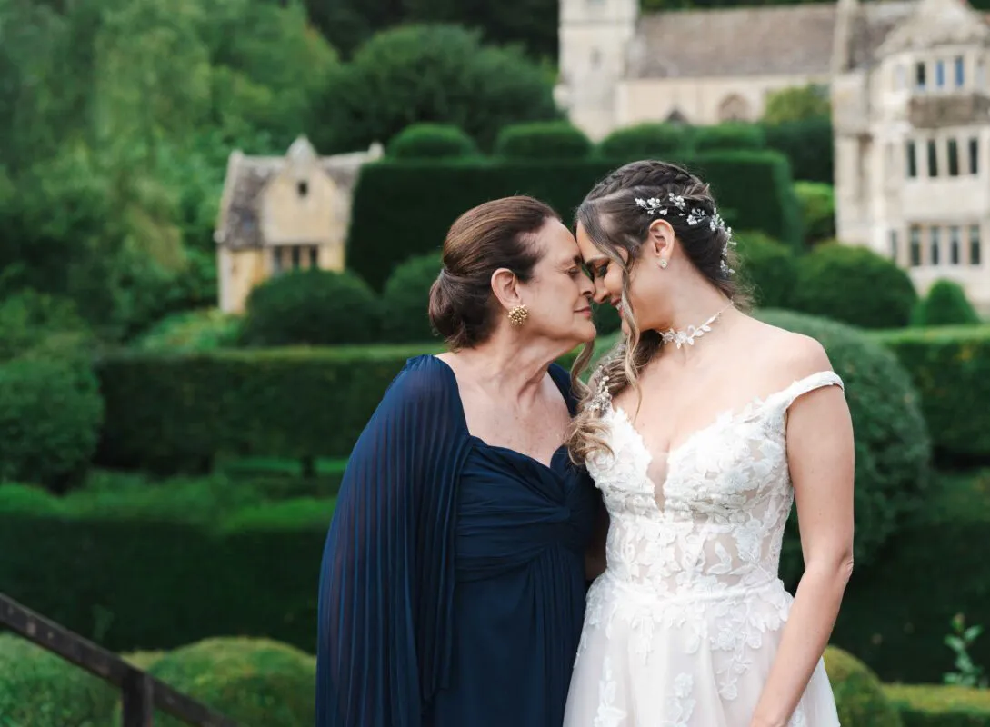 A bride and her mother share a tender nose-to-nose moment in a manicured country house garden, with the mother wearing an elegant navy blue gown and the bride wearing a lace off-the-shoulder wedding dress.