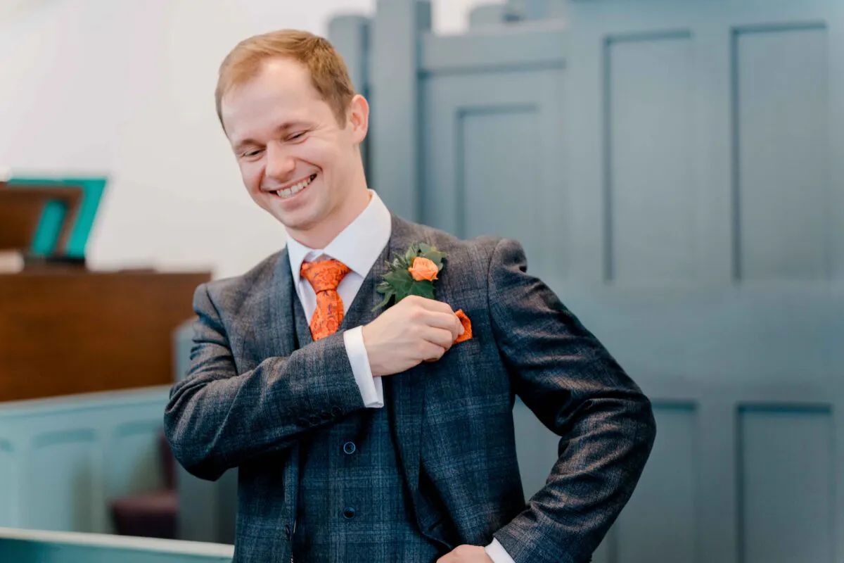 A smiling groom adjusts his orange pocket square while wearing a checked grey three-piece suit with a matching orange tie and boutonniere.