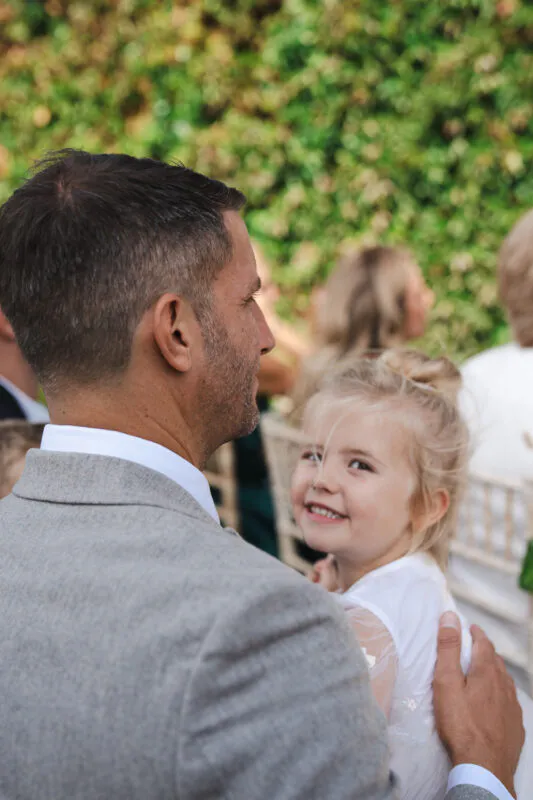 A man wearing a grey suit and white shirt holds a smiling young girl wearing a white dress against a backdrop of green foliage.