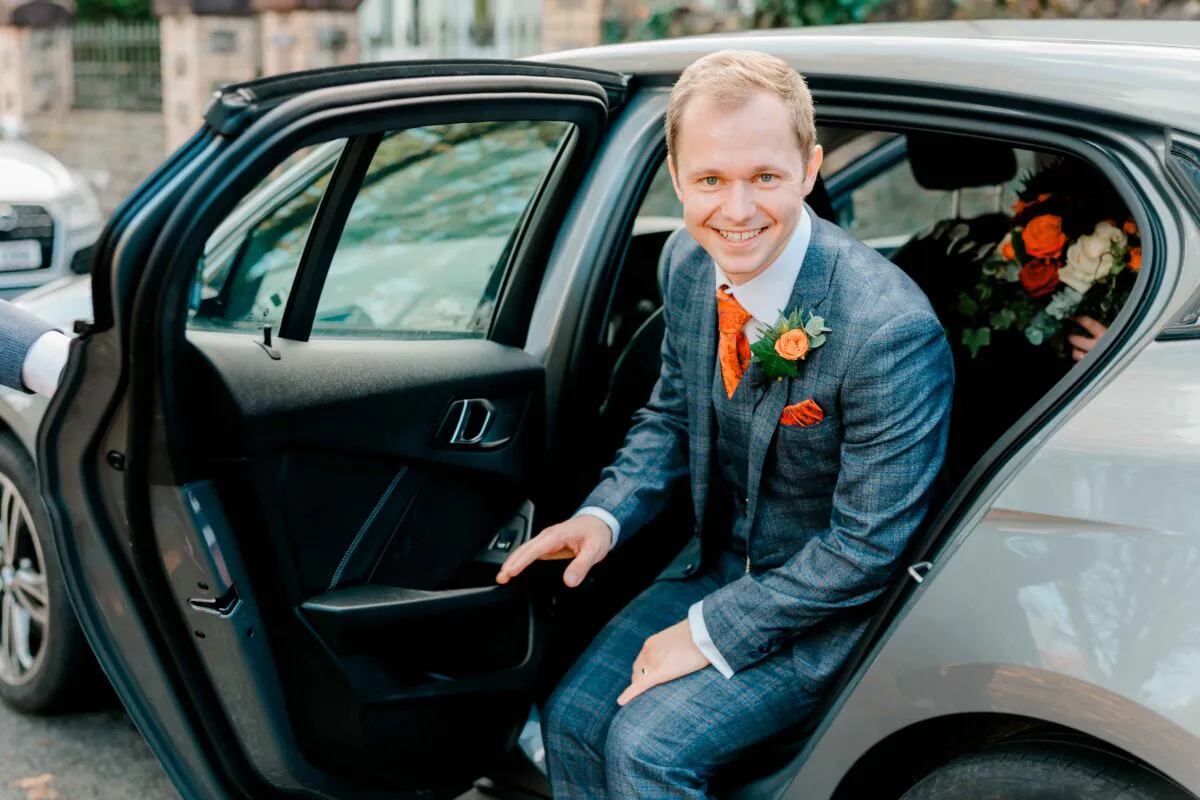 A smiling groom in a grey checked three-piece suit steps out of a car, wearing a patterned orange tie and matching boutonniere.