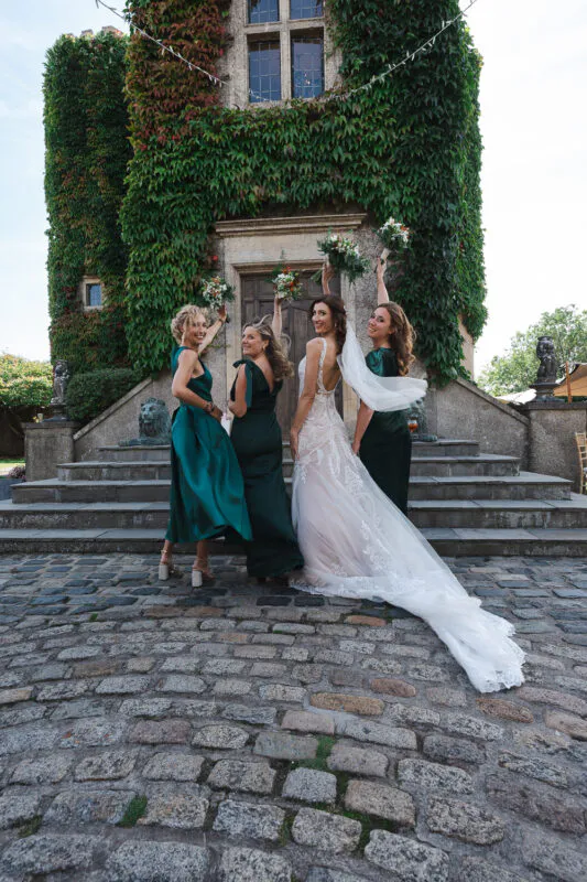 The bride and her bridesmaids in green dresses raise their bouquets in celebration outside a historic ivy-clad castle entrance.