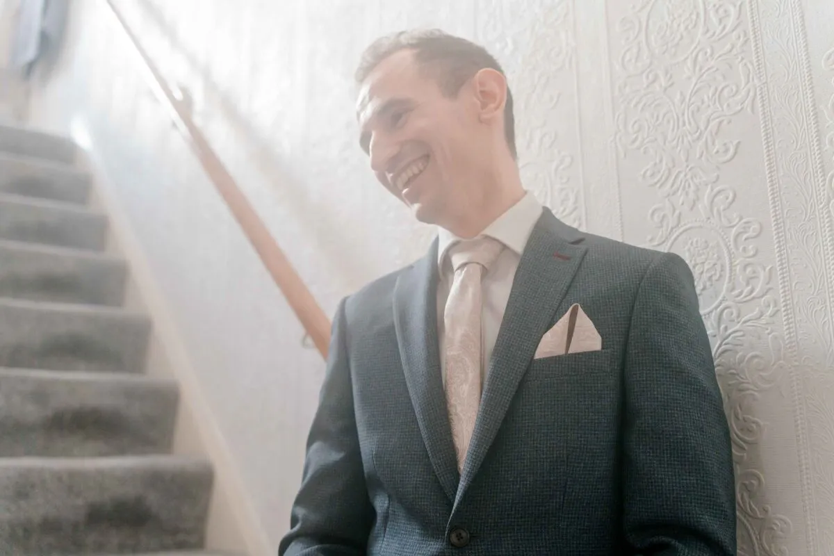 A smiling groom in a grey suit and patterned tie stands near a staircase, caught in a joyful candid moment.