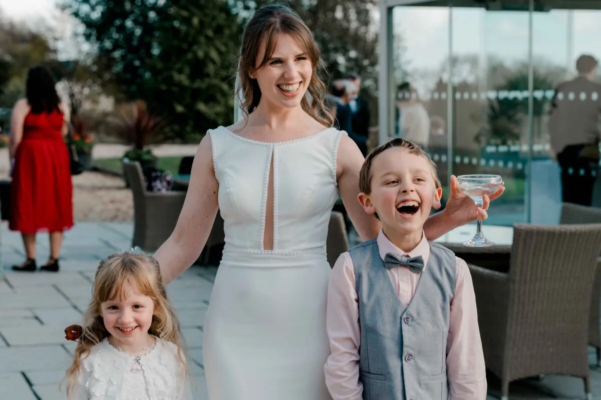 A joyful bride smiles broadly while standing outside on a patio with a laughing page boy holding a champagne coupe and a smiling flower girl.