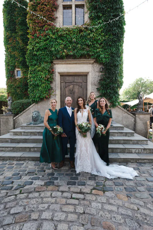 A newlywed couple and their bridesmaids pose on a cobbled courtyard in front of a historic castle tower covered in lush green ivy.
