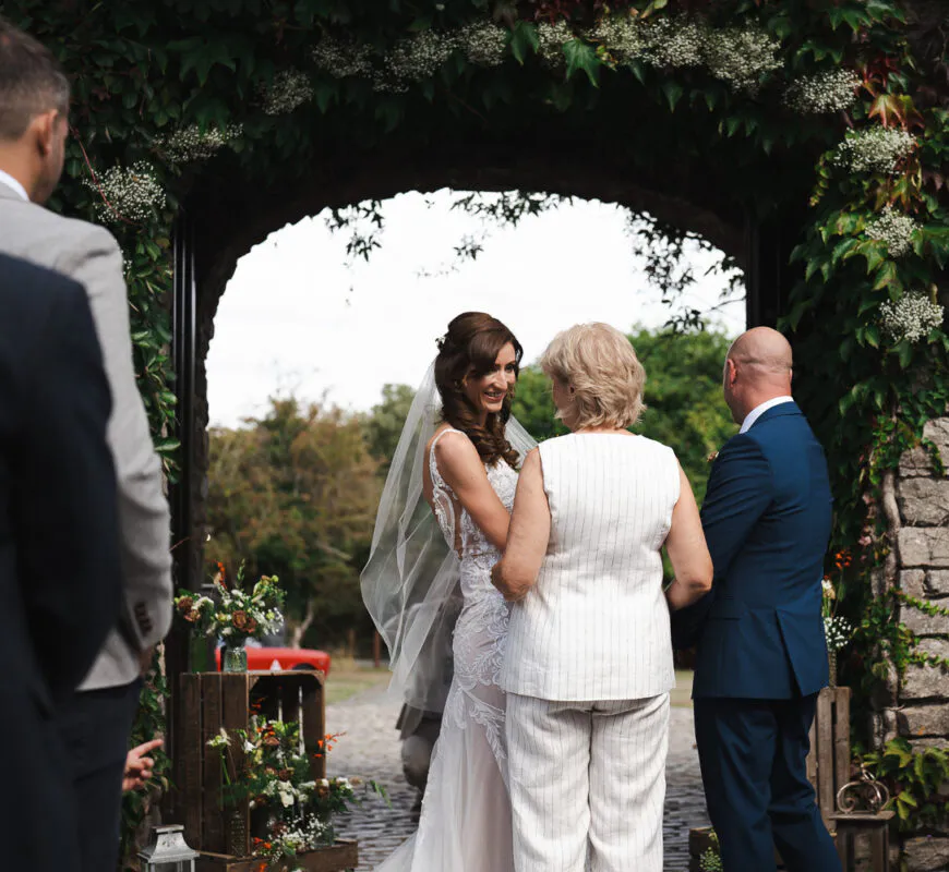 A smiling bride in a lace wedding dress and veil stands near an ivy-covered stone archway while interacting with guests during an outdoor ceremony.