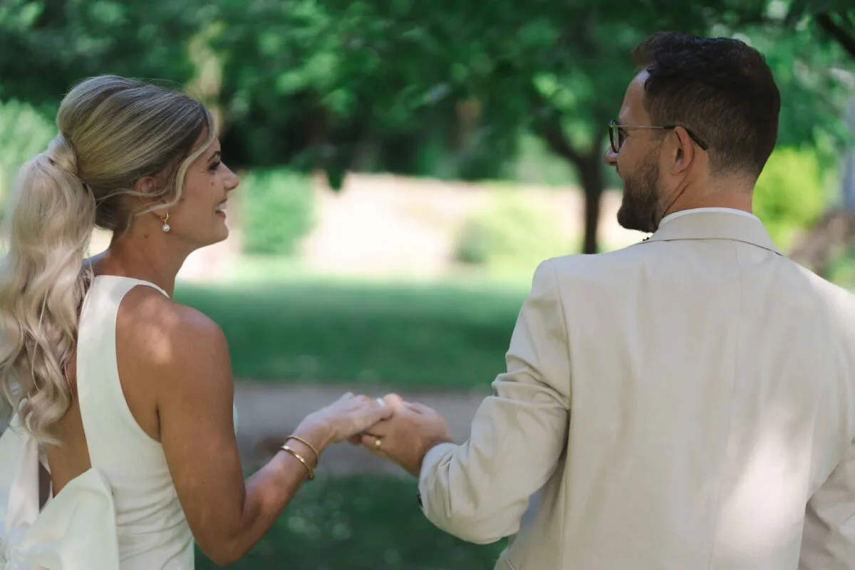 A bride in a modern sleeveless gown with a ponytail and a groom in a beige suit hold hands and share a tender glance outdoors.