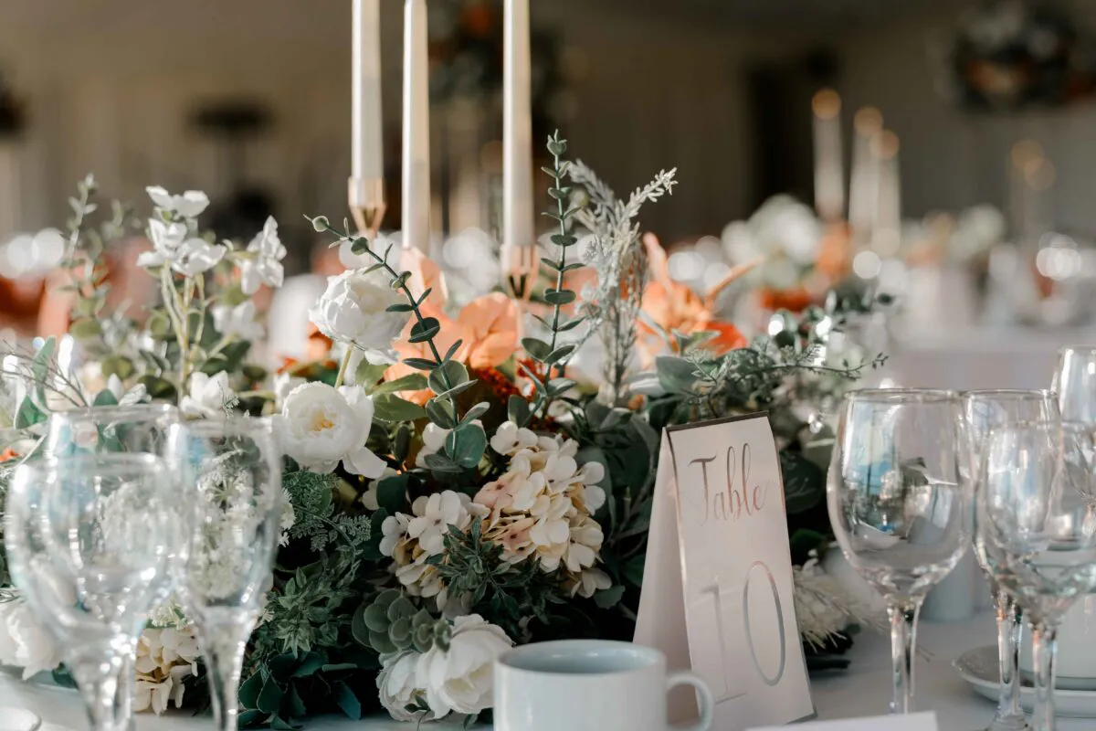 An elegant wedding tablescape features a lush floral centrepiece of white roses, hydrangeas, and eucalyptus, accompanied by taper candles and a table number sign.