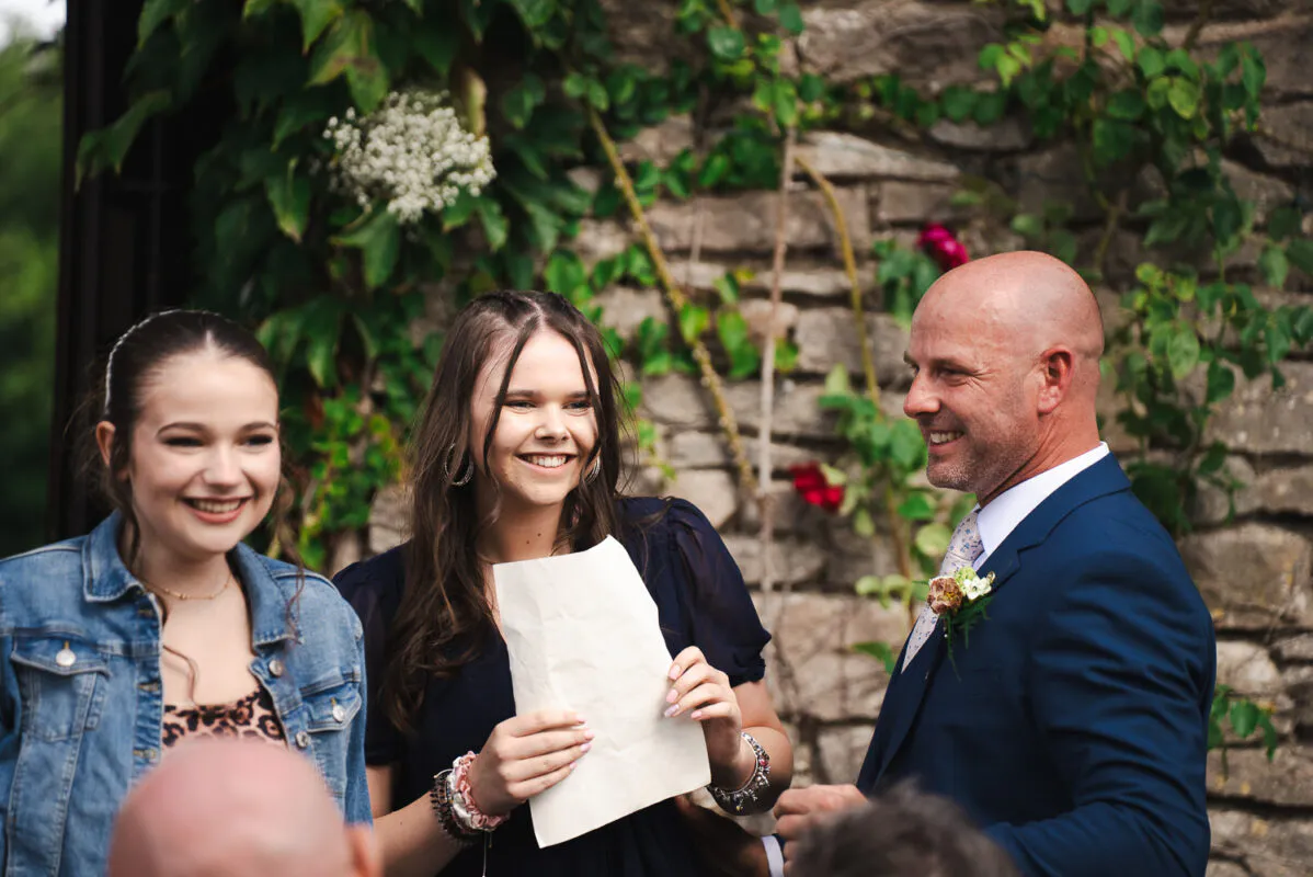 A smiling groom in a navy suit looks on as guests laugh while reading a letter outside against a rustic stone wall.