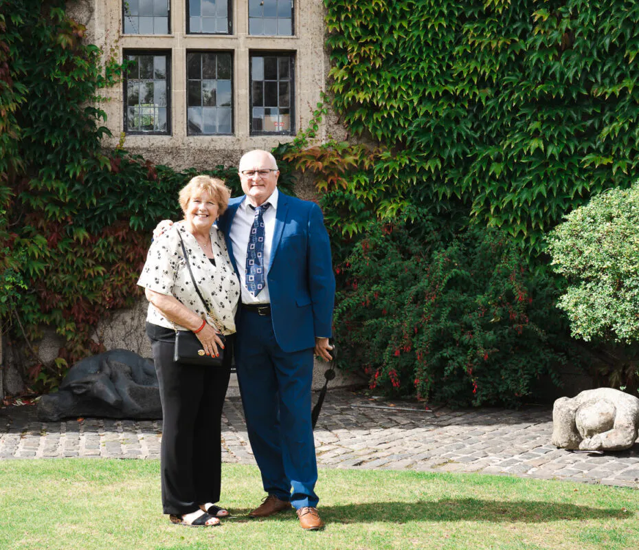 An older couple smiling together on a cobbled courtyard in front of an ivy covered stone building during a wedding reception.
