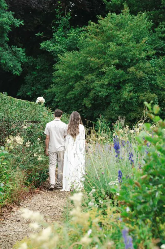 A newlywed couple walks away together along a gravel pathway surrounded by lush green foliage and vibrant wildflowers in a garden setting.