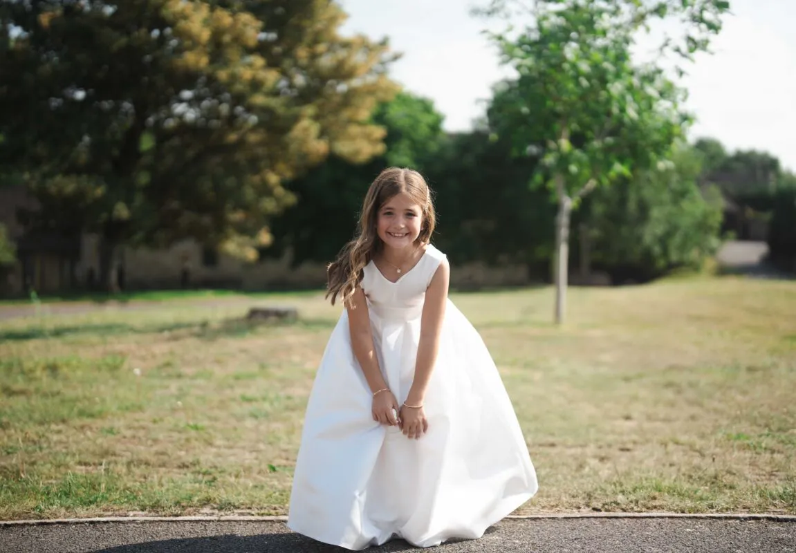A smiling young flower girl wearing a white dress stands outside on a grassy path during a summer wedding.