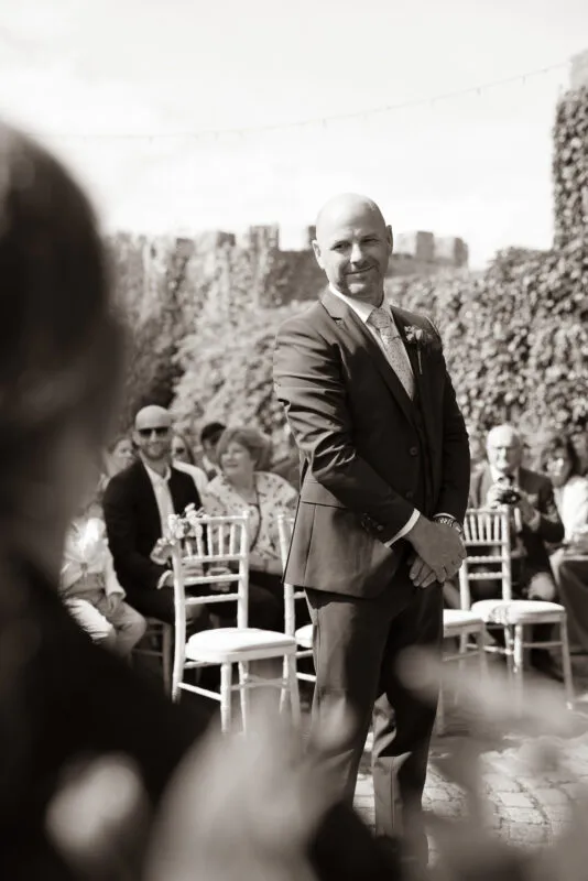 An emotional groom smiling happily as he stands at the altar during an outdoor wedding ceremony, viewed from over the shoulder of the bride.