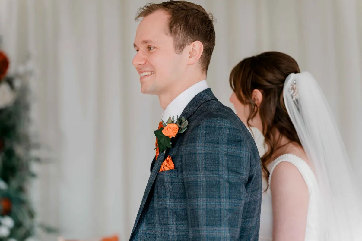 A smiling groom wearing a blue checked suit and orange buttonhole stands back-to-back with his bride during their wedding first look.