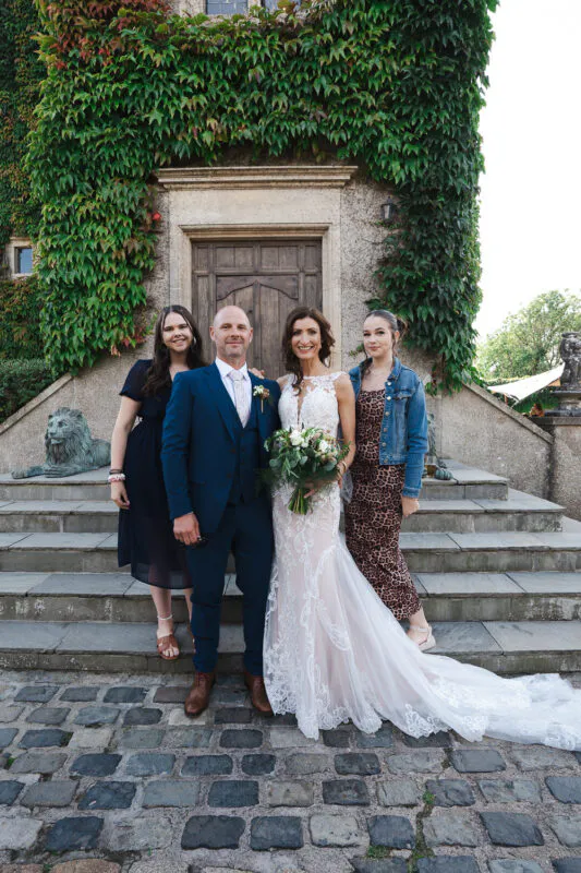 The bride and groom pose on stone steps in front of an ivy covered stone building with two wedding guests standing beside them.