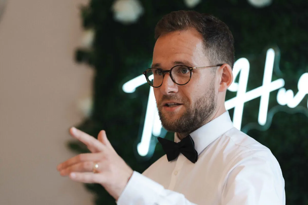 A groom wearing glasses and a crisp white shirt with a black bow tie gestures while speaking in front of a neon sign and green floral wall.