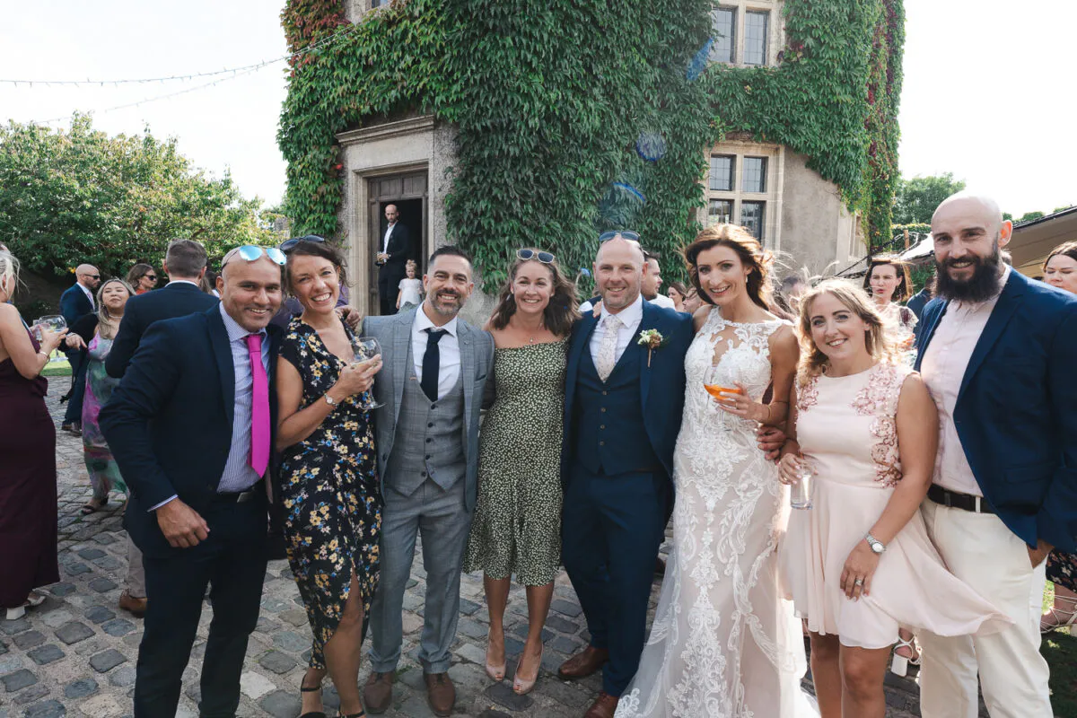 A group of smiling wedding guests and the newlyweds pose together in a cobbled courtyard in front of an ivy-covered historic stone building during a sunny outdoor drinks reception.