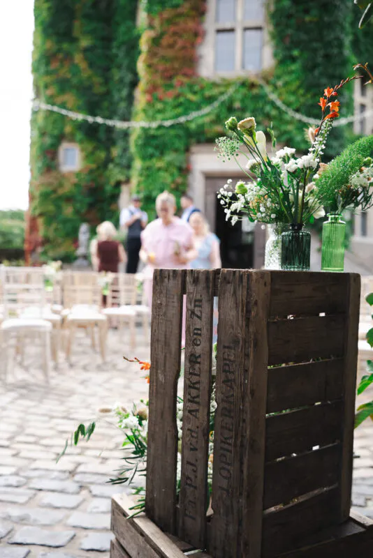 A rustic wooden crate adorned with bud vases filled with wildflowers stands in a cobbled courtyard with wedding guests blurred in the background.
