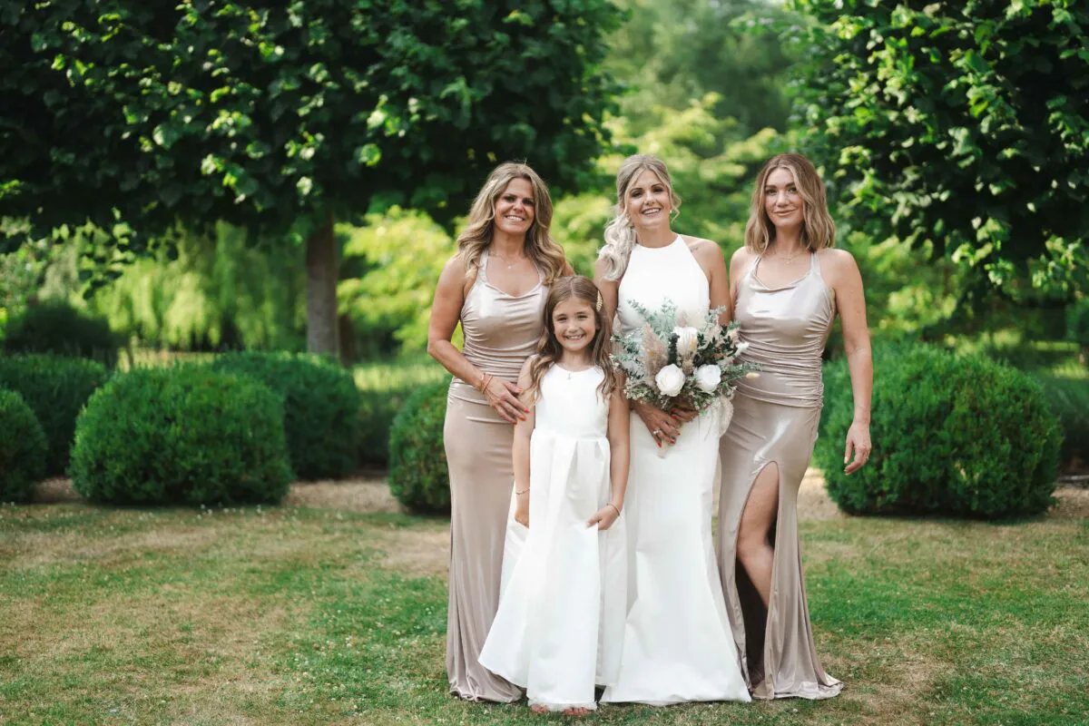 A smiling bride holding a bouquet of white roses and foliage poses outdoors in a garden alongside two bridesmaids in champagne satin dresses and a young flower girl.