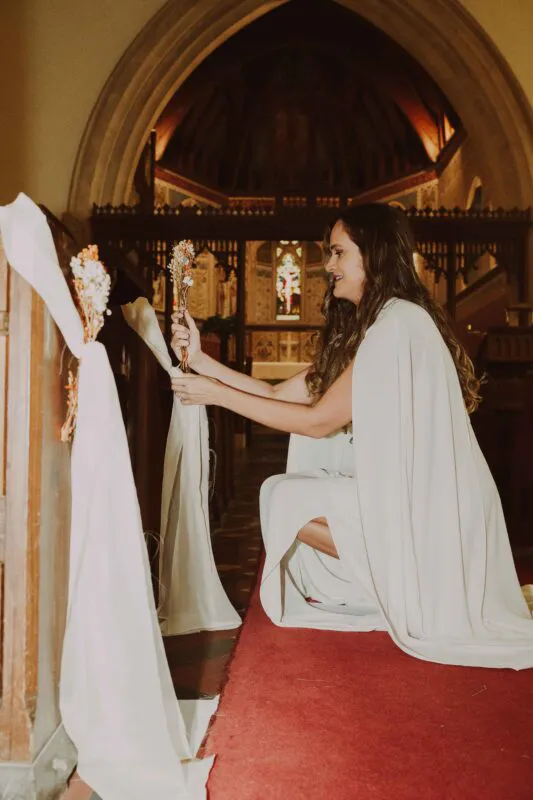 A smiling bride wearing a white cape and jumpsuit kneels inside a traditional church, arranging pew decorations adorned with dried flowers.